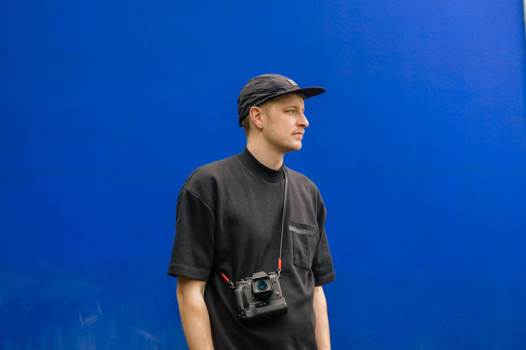 Low angle view of young man wearing cap looking away while standing against blue wall