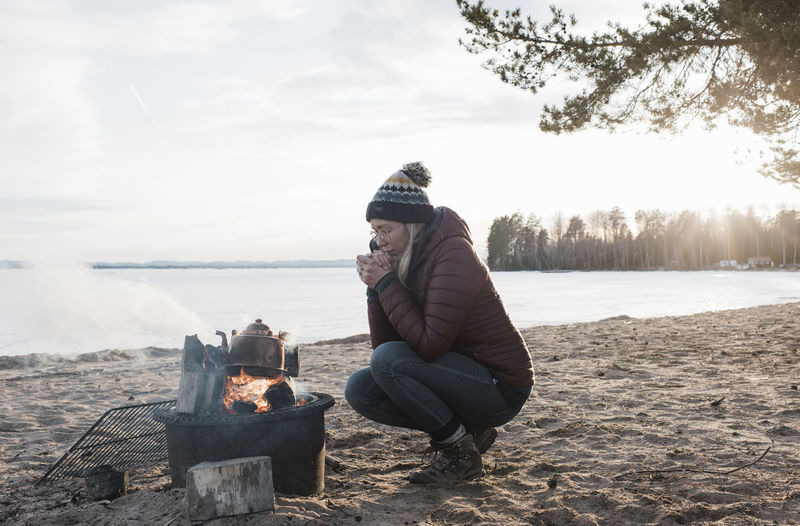 Woman warming up sat next to a camp fire on an empty beach in sweden