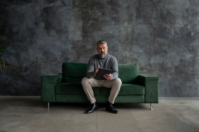 Portrait of handsome senior man sitting on green couch, using smartphone in stylish loft apartment