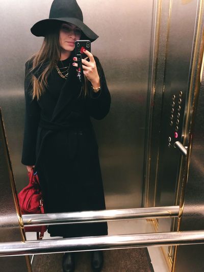 Young woman taking selfie with smart phone reflecting on mirror in elevator