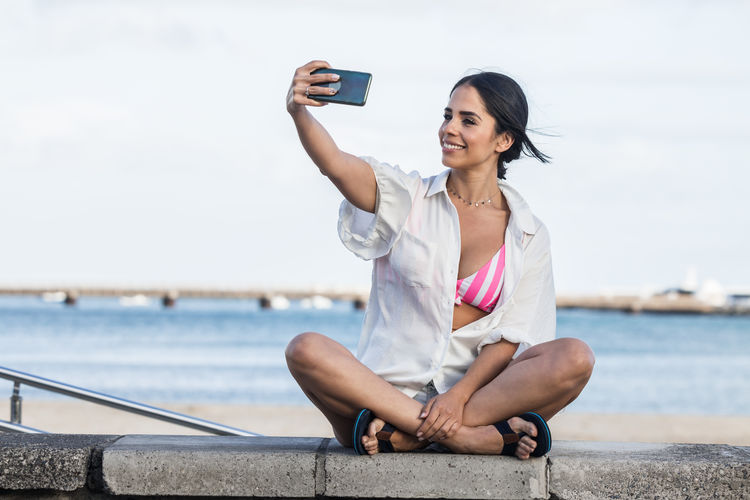 Full length of young woman using mobile phone while sitting on shore