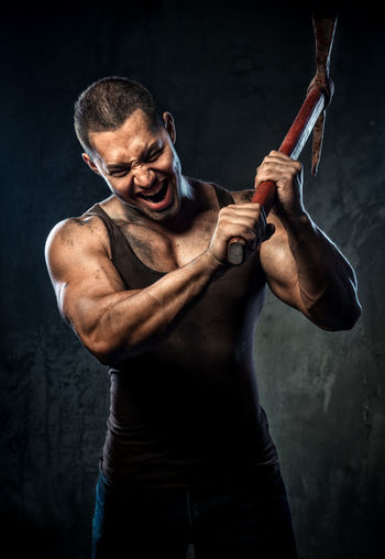Aggressive muscular man holding pick axe against wall
