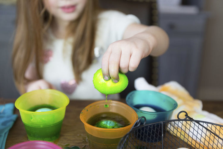 Midsection of girl dipping easter egg in dyed water on table at home