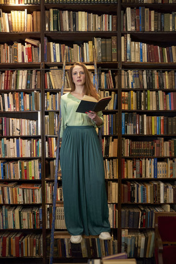 Young woman holding book while standing on ladder in library