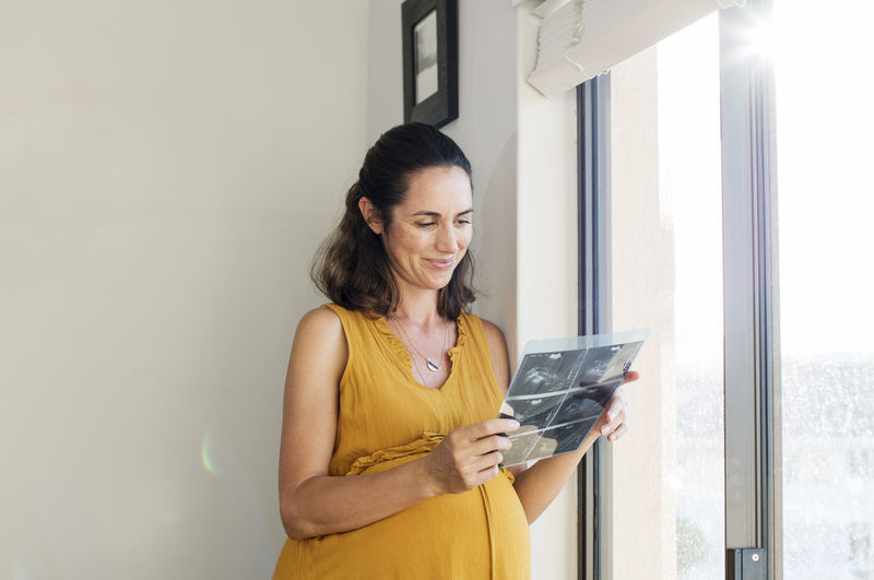 Smiling pregnant woman looking at x-ray while standing by window