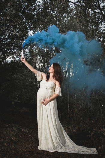 Pregnant woman holding distress flare while standing against trees in forest