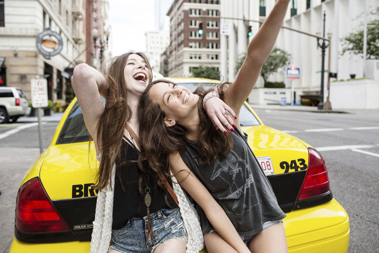 Cheerful female friends sitting on yellow taxi on road in city