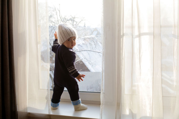 Child in a warm blue jumpsuit is on window in winter white knit hat