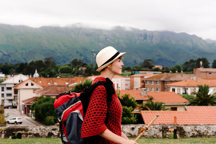 Side view of happy female backpacker with stick standing on hill and exploring old town located against mountains during pilgrimage trip through spain