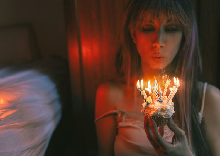 Young woman blowing burning candles on birthday cupcake at home