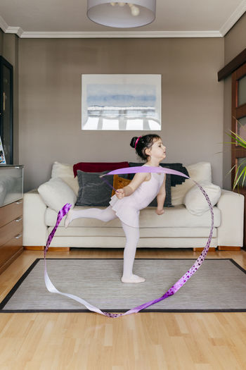 Side view of cute little girl in leotard and tights spinning ribbon and dancing during rhythmic gymnastic training in cozy living room at home