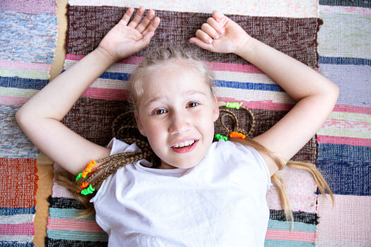 Portrait of a beautiful smiling girl with pigtails smiling, lying on a woven rug with her arms up.