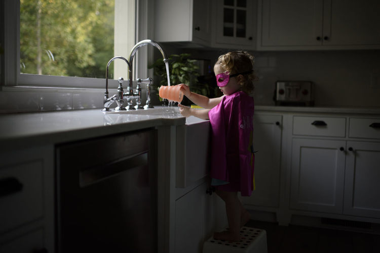 Side view of girl wearing superhero costume washing hands in kitchen sink while standing at home