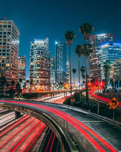 Light trails on city street by buildings against sky at night