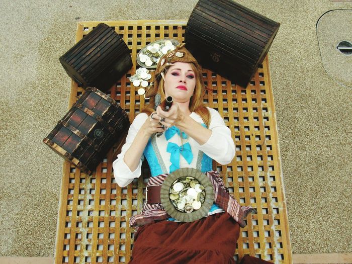High angle portrait of female pirate pointing gun while lying amidst treasure chest