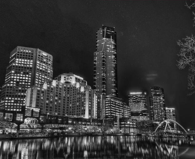 Illuminated modern buildings by river against sky at night