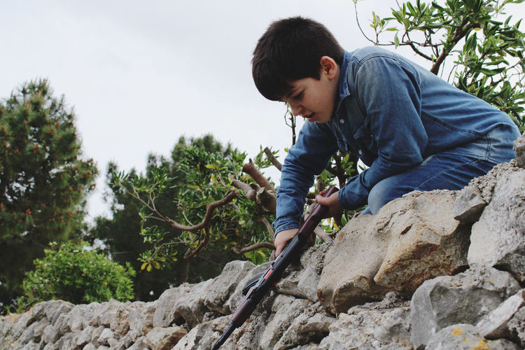 Low angle view of boy with toy gun kneeling on stone wall