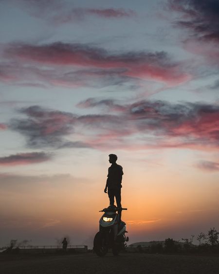 Silhouette man standing on motor scooter against sky during sunset