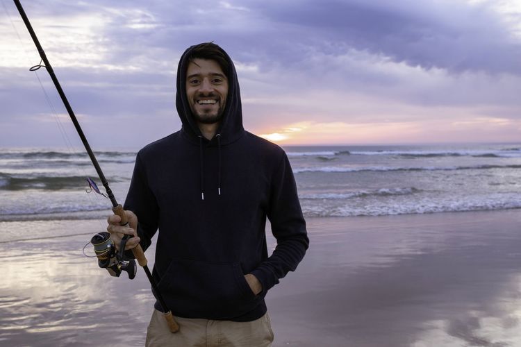 Portrait of man holding fishing rod standing at beach against sky