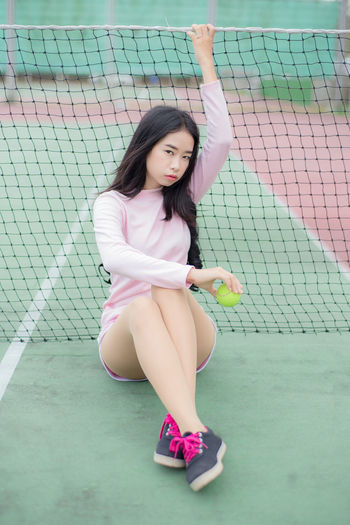 Full length portrait of young woman holding ball while sitting on court