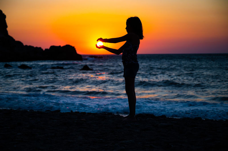 Side view of woman jumping at beach against sky during sunset