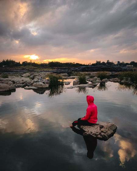 Rear view of man sitting on lake against sky during sunset