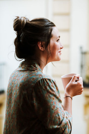 Side view of happy female brunette in casual flowery shirt smiling away while having hot drink against blurred interior of light room