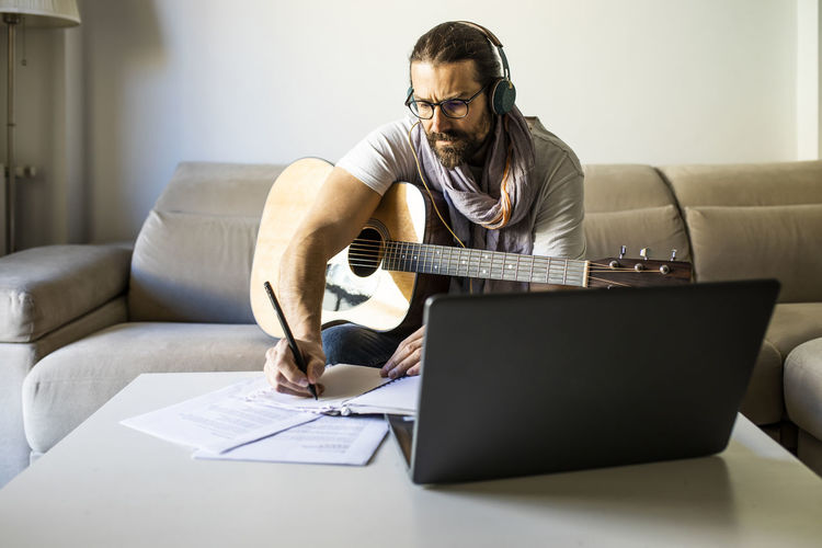 Bearded male musician in eyeglasses sitting on couch with guitar and writing notes on table while using headphones and laptop