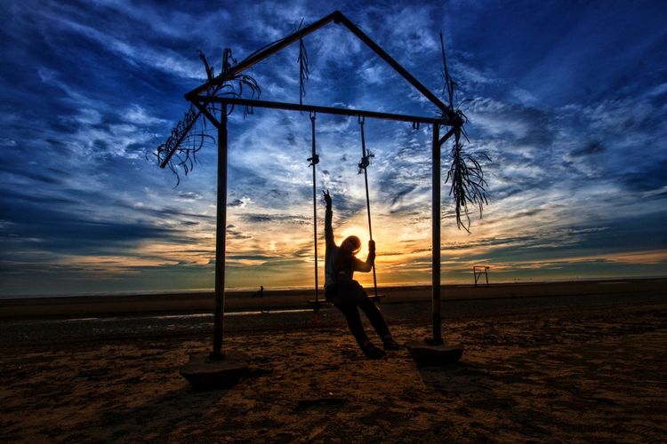 Silhouette woman standing on swing against sky during sunset