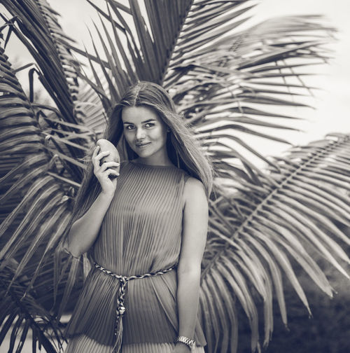 Beautiful woman holding mango while standing against palm leaves