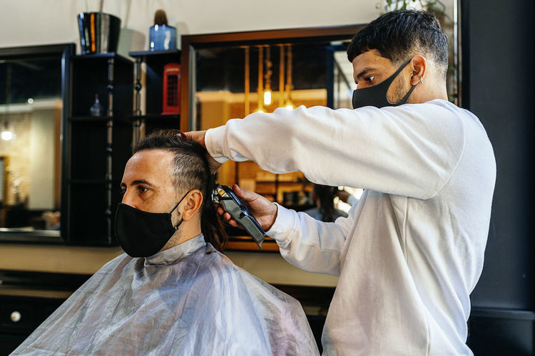 Side view of focused male hairdresser using trimmer and making hairdo for client in modern barbershop