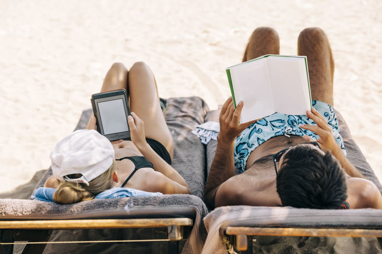 High angle view of man reading book while woman using digital tablet on lounge chairs at beach