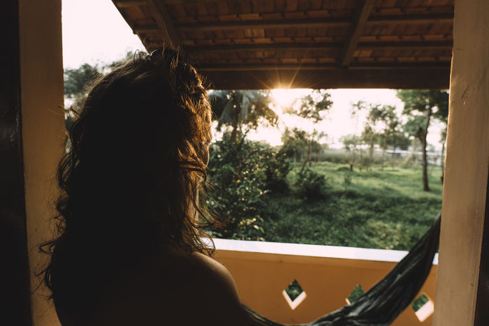 Rear view of woman standing by window during sunset
