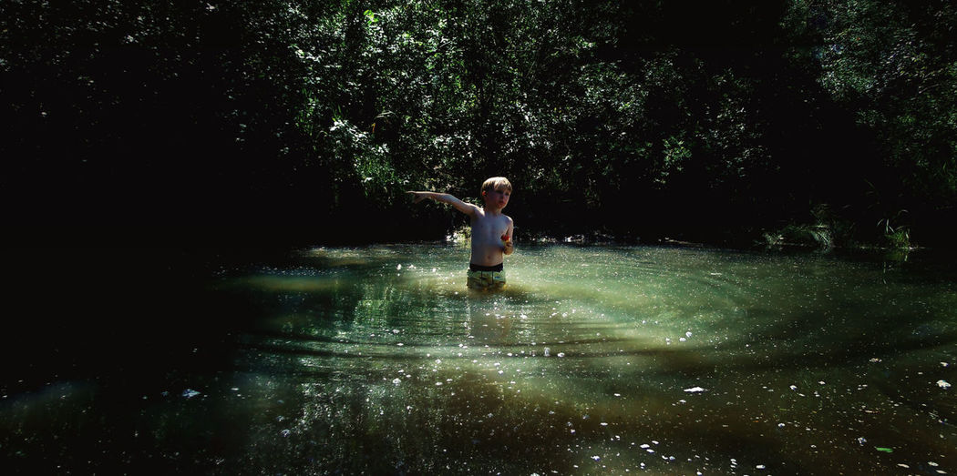 Shirtless boy standing in lake against trees
