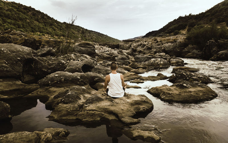 Man from behind practicing yoga on a rock in the river and reflection in the water