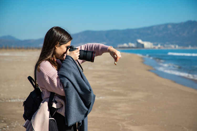 Side view of young woman photographing while standing at beach