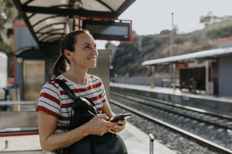 Smiling woman with smart phone standing at railroad station
