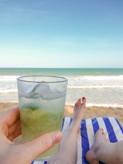 Cropped image of hand holding drink at beach against sky