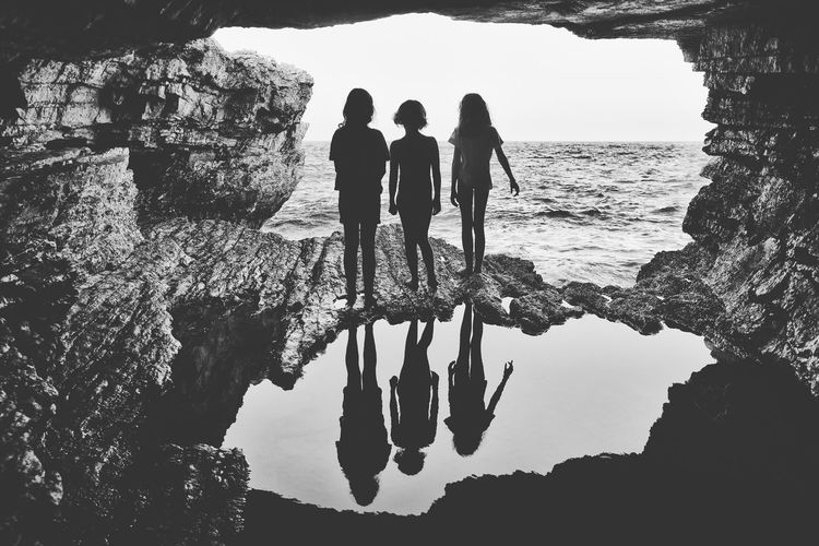 Rear view of silhouette girls standing on rock by sea