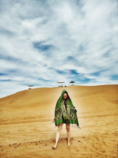 Full length of woman wrapped in shawl standing on sand at beach against cloudy sky