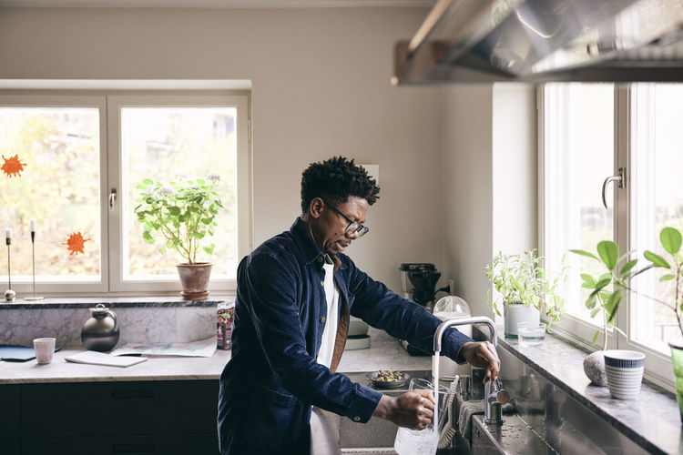 Side view of mature man filling pitcher from faucet in kitchen at home