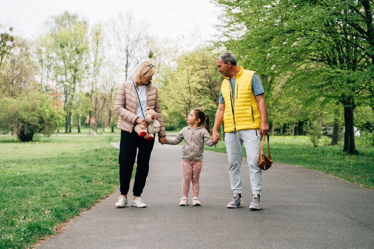 Mature grandparents in casual clothes holding hands of adorable happy little girl while walking together on path in green park
