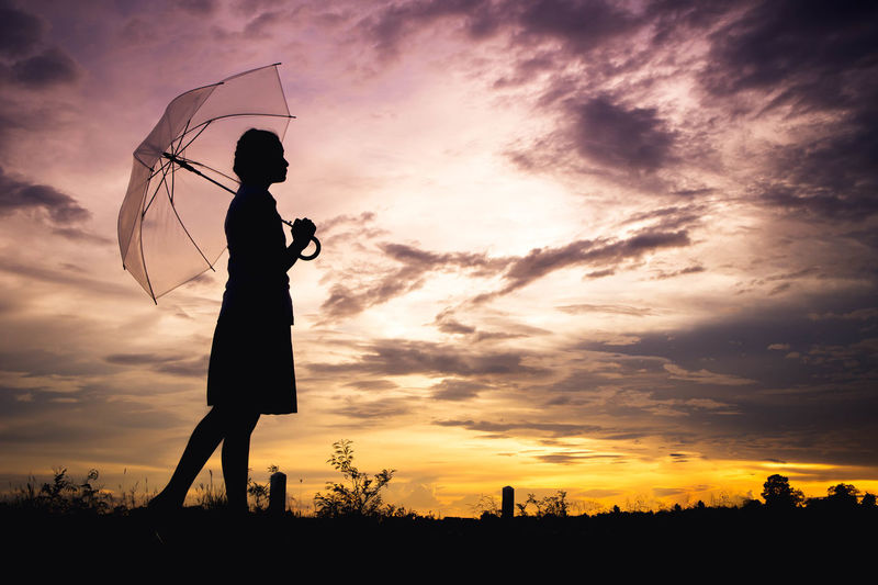 Silhouette mid adult woman carrying umbrella while standing on field against sky during sunset
