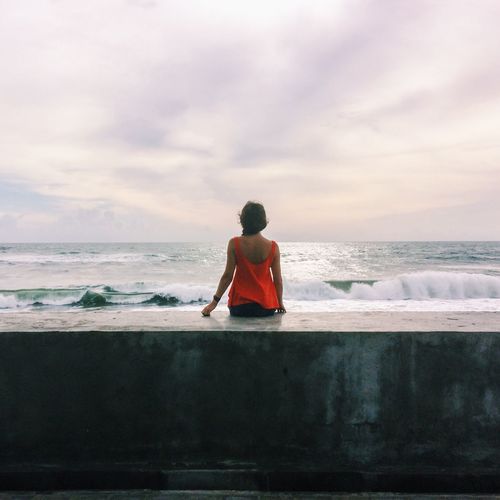 Rear view of woman sitting on retaining wall by sea against cloudy sky