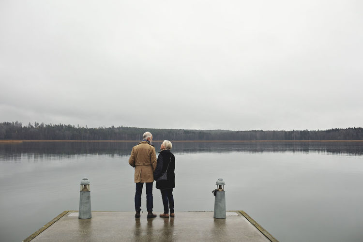 Rear view of senior couple talking while standing by lake against clear sky