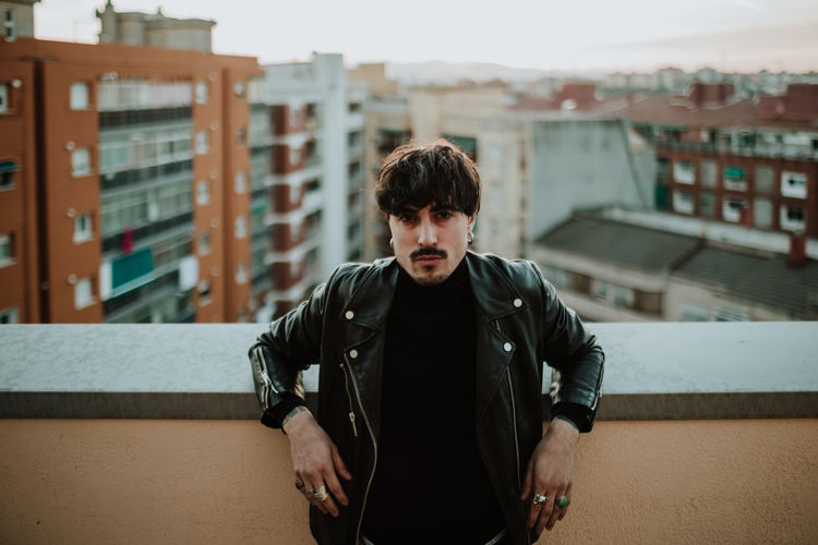 Serious man in leather jacket standing on rooftop and leaning on fencing and confidently looking at camera