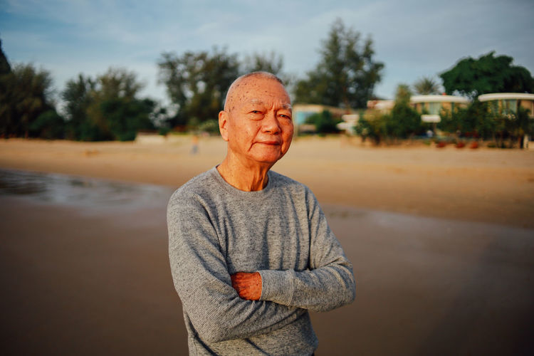 Portrait of senior man with arms crossed standing at beach against sky during sunset