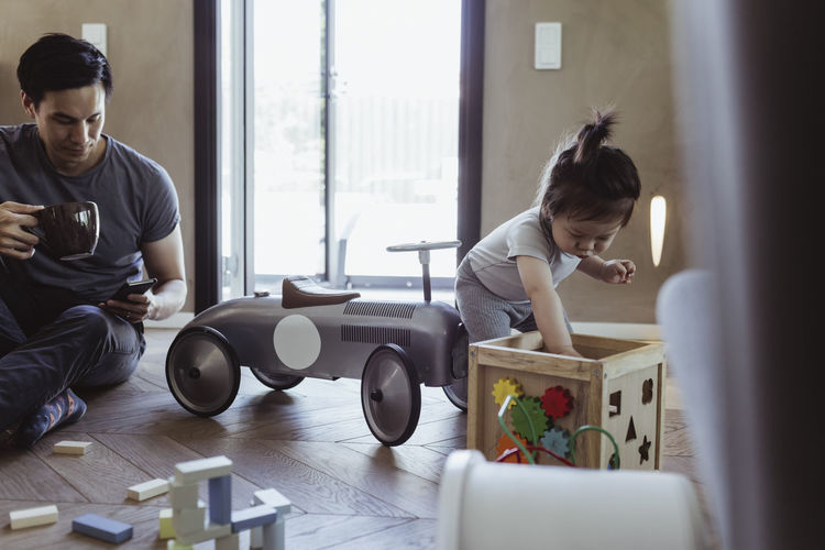 Male toddler playing with puzzle toy while father using smart phone in living room
