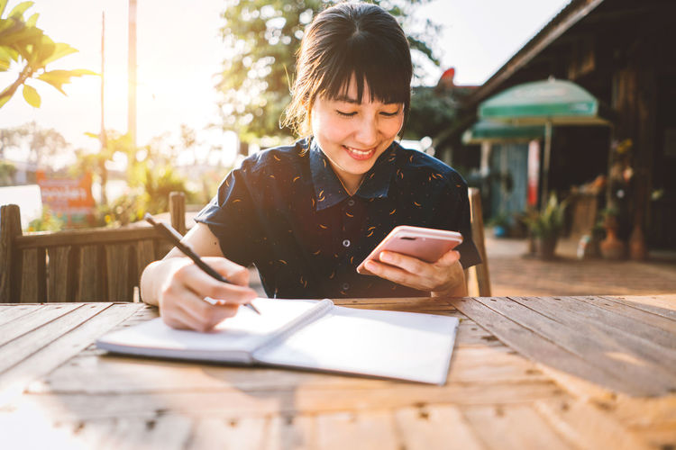 Woman using mobile phone while writing in book on table