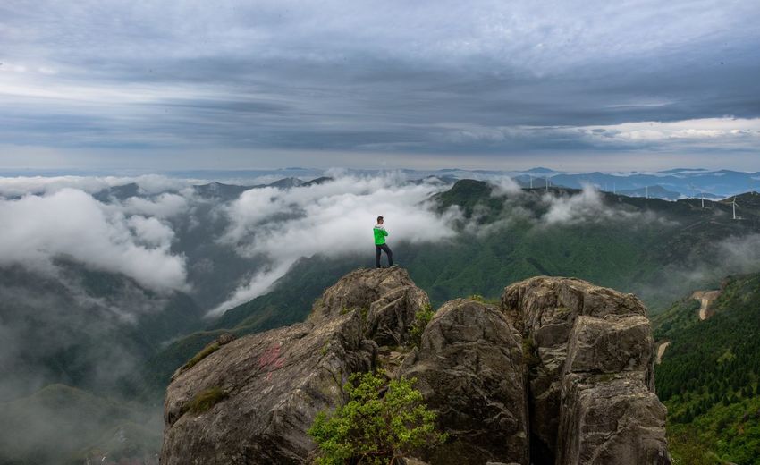 Man standing on cliff overlooking mountains and wind turbine